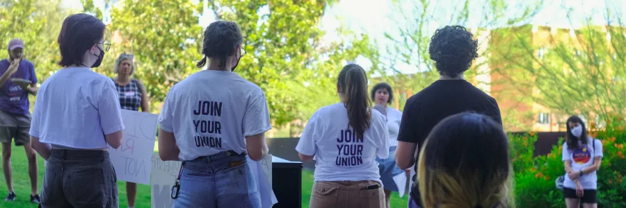 The backs of several people standing on stage during a rally/press conference. Two of them wear white tshirts that read "Join Your Union" in blue.