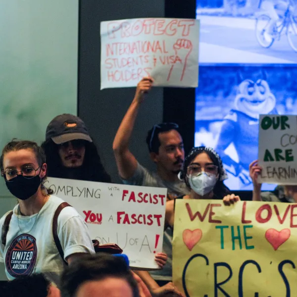 Multiple people holding signs in protest at an Arizona Board of Regents meeting. One person on the left wears a UCWAZ shirt.