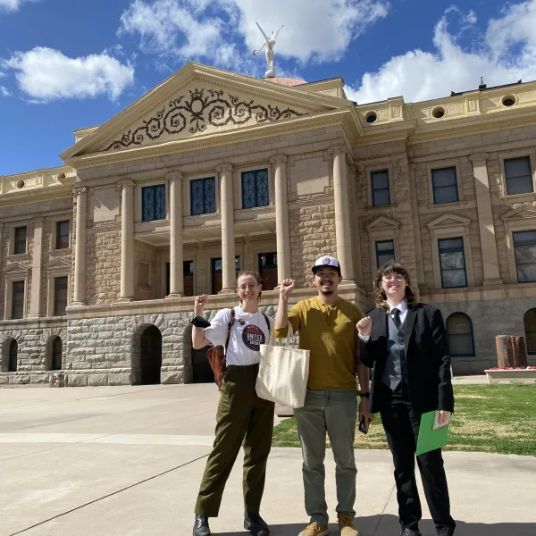 Three people stand in front of the state capitol building in Phoenix, AZ. One wears a UCWAZ hat and one wears a UCWAZ shirt.